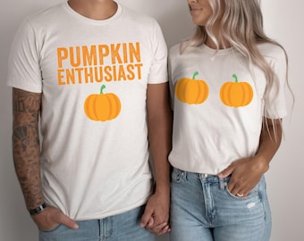 May include: Two white t-shirts with pumpkin graphics. One shirt says "Pumpkin Enthusiast" and has a single orange pumpkin. The other shirt has two orange pumpkins.