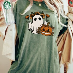 K&ouml;nnte beinhalten: Olivgr&uuml;nes T-Shirt mit einem Halloween-Motiv. Das Design zeigt einen wei&szlig;en Geist mit Sonnenblumenkranz, orangefarbenen Blumen und einem K&uuml;rbis. Das T-Shirt hat eine lockere Passform.