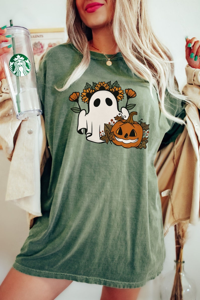 K&ouml;nnte beinhalten: Olivgr&uuml;nes T-Shirt mit einem Halloween-Motiv. Das Design zeigt einen wei&szlig;en Geist mit Sonnenblumenkranz, orangefarbenen Blumen und einem K&uuml;rbis. Das T-Shirt hat eine lockere Passform.