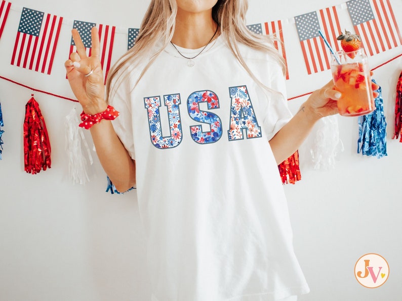 Op de afbeelding: Een wit T-shirt met een bloemenprintontwerp dat "USA" spelt in rood, wit en blauw. Het shirt wordt gedragen door een persoon die een glas met rode vloeistof vasthoudt.