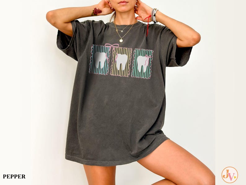 May include: Dark gray oversized t-shirt featuring a graphic of three teeth with colorful striped backgrounds and bows. The shirt has short sleeves and a relaxed fit. The word "PEPPER" is printed at the bottom left.