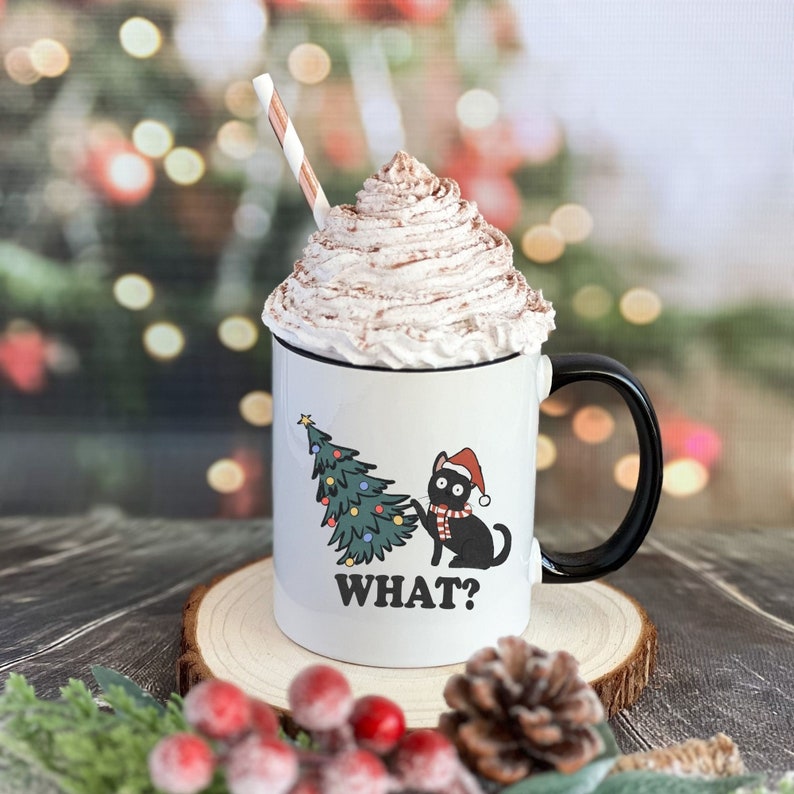 May include: White ceramic mug with a black cat wearing a Santa hat and a Christmas tree design. The mug has the text "WHAT?" printed on it. The mug is filled with whipped cream and a straw.