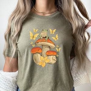 May include: A green frog sitting on a mushroom with orange and brown accents. The mushroom is surrounded by yellow flowers and butterflies. The image is printed on a green t-shirt.