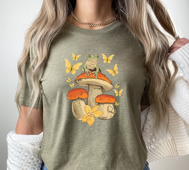May include: A green frog sitting on a mushroom with orange and brown accents. The mushroom is surrounded by yellow flowers and butterflies. The image is printed on a green t-shirt.
