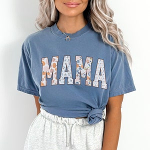 May include: A blue t-shirt with the word "MAMA" printed in large, white letters. The letters are decorated with a floral pattern in shades of pink, orange, and blue.