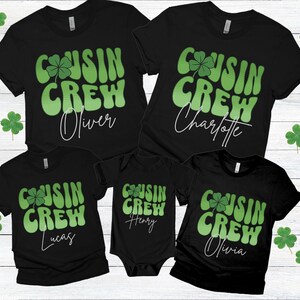 May include: Black t-shirts and a onesie with the text "COUSIN CREW" in green, featuring a shamrock. Names Oliver, Charlotte, Lucas, Henry, and Olivia are printed below the text. The shirts are arranged on a light wood background.
