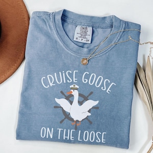 Puede incluir: Una camiseta azul claro con un gr&aacute;fico blanco de un ganso con un sombrero de capit&aacute;n y un salvavidas. El texto "Cruise Goose on the Loose" est&aacute; impreso en blanco en la camiseta.