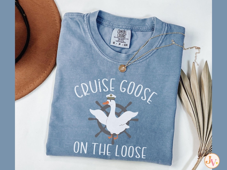 Puede incluir: Una camiseta azul claro con un gr&aacute;fico blanco de un ganso con un sombrero de capit&aacute;n y un salvavidas. El texto "Cruise Goose on the Loose" est&aacute; impreso en blanco en la camiseta.