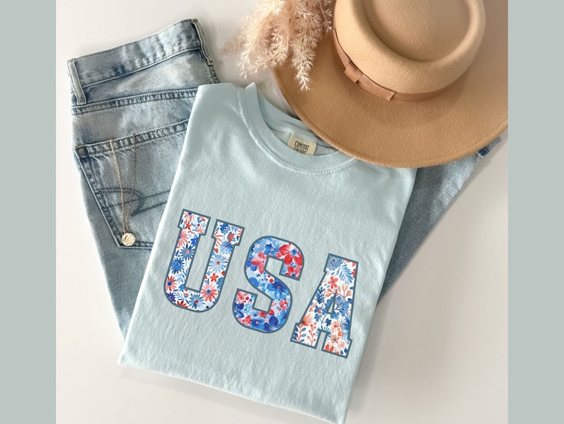 Op de afbeelding: Een lichtblauw T-shirt met het woord "USA" erop gedrukt. De letters zijn gemaakt van een bloemenpatroon in rood, wit en blauw.