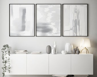 May include: Three abstract paintings in black frames with gray and white brushstrokes, displayed on a white wall above a white cabinet with a gray vase and other decorative items.