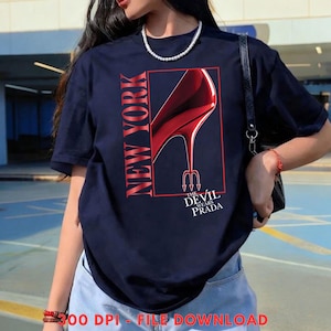 May include: Navy blue t-shirt with a red graphic of a high-heeled shoe and the text "NEW YORK" and "THE DEVIL WEARS PRADA". The shoe has a devil's pitchfork heel. The shirt is a casual fashion item.