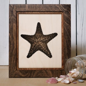 May include: A framed wood cutout of a starfish in a dark brown frame. The frame is sitting on a wooden surface with a glass jar of seashells spilling out.