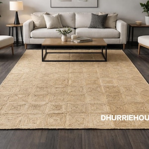 May include: A rectangular jute rug with a geometric square pattern. The rug is a natural beige color and covers a dark wood floor. The rug is in a living room setting with a sofa, chairs, and a coffee table.