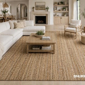 Puede incluir: Una alfombra de yute rectangular grande de color marrón claro natural. La alfombra está en un salón con un sofá blanco, una mesa de centro de madera y un sillón. La alfombra tiene un aspecto texturizado y tejido.