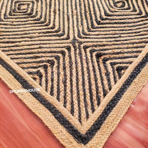 Op de afbeelding: Een rechthoekig vloerkleed gemaakt van gevlochten natuurlijke jute en zwarte vezels, met een geometrisch patroon van concentrische vierkanten. De tekst DHURRIEHOUSE staat in het wit gedrukt in de linkerbenedenhoek van het oppervlak.