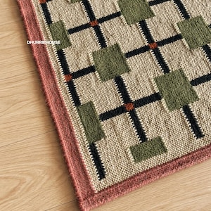 May include: A woven rug featuring a grid pattern of black lines on a beige background with alternating green squares and small orange accents at the intersections. The rug has a solid dusty pink border and the text DHURRIEHOUSE printed on it.