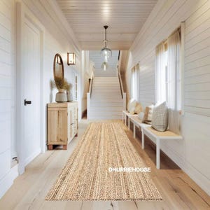 May include: A hallway with a natural jute runner rug. The rug has a rectangular shape with vertical woven stripes. The hallway features white walls, a wooden floor, a staircase, and a bench with cushions. The text "DHURRIEHOUSE" is visible on the rug.