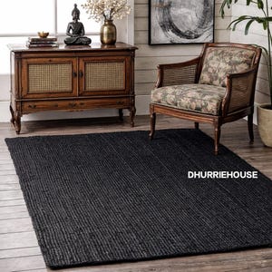 以下が含まれることがあります： 木製の床に置かれた、テクスチャ加工されたリブ加工の表面を持つ、大きな長方形の黒いラグ。画像には、木製のキャビネット、模様入りのアームチェア、仏像も含まれています。