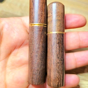 May include: Two wooden cylindrical containers with a dark brown finish and a gold band around the middle. The containers are held in a hand, showcasing their smooth, polished texture and natural wood grain.