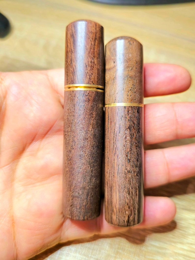 May include: Two wooden cylindrical containers with a dark brown finish and a gold band around the middle. The containers are held in a hand, showcasing their smooth, polished texture and natural wood grain.