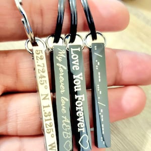 Peut inclure: Quatre porte-clés rectangulaires avec des messages gravés. L'un affiche des coordonnées, un autre dit "My forever love A&B", un troisième dit "Love You Forever", et le dernier a du code Morse. Les porte-clés sont tenus dans une main.