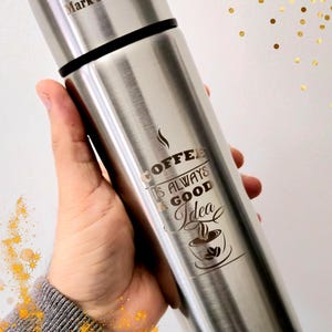 Op de afbeelding: Een roestvrijstalen koffiethermoskan met een zwarte band en een zilveren deksel. De thermoskan heeft de tekst "Mark's coffee" bovenaan en de zin "Coffee is always a good idea" met een koffiekopje.