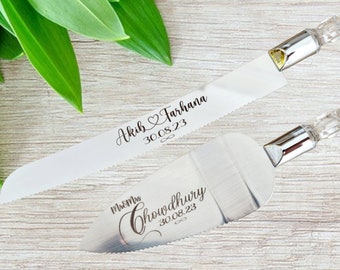 May include: A wedding cake knife and server set. The knife is white with a serrated edge and the server is silver. Both have clear crystal handles. The knife is engraved with "Akib & Farhana 30.08.23". The server is engraved with "Mr & Mrs Chowdhury 30.08.23".