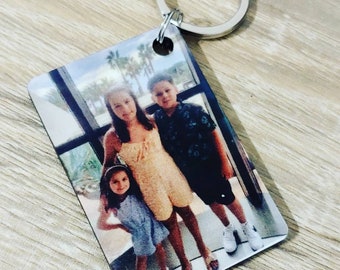 May include: Rectangular keychain with a printed photograph of three people. The photo shows two girls and a boy standing in front of a window. The keychain has a silver metal ring attached to the top.