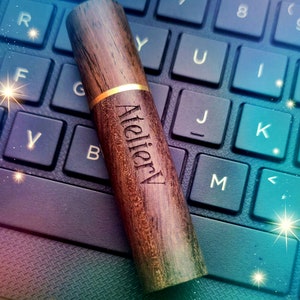 May include: A wooden pen with the text "AtelierV" engraved on the side. The pen has a dark brown finish with a gold band and is resting on a black laptop keyboard.