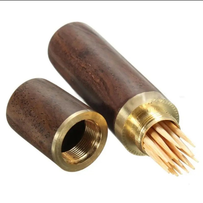 May include: A wooden toothpick holder with a brass-colored metal rim and screw-on cap. The holder is open, revealing a bundle of wooden toothpicks. The wood has a dark brown grain.