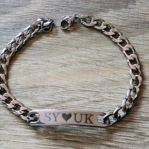 Puede incluir: Una pulsera de cadena de color plateado con una placa rectangular. La placa está grabada con las letras "SY", un símbolo de corazón y "UK". La pulsera está hecha de un material metálico brillante y se muestra sobre una superficie de madera.