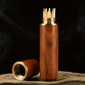 May include: A wooden toothpick holder with a brass-colored metal top and bottom. The holder is cylindrical and made of dark brown wood. The lid is off, revealing a bundle of toothpicks. The background is dark.