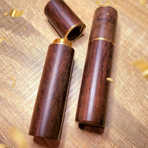 May include: Three cylindrical wooden containers with brass accents. One container is open, revealing the threaded interior. The wood grain is visible, and the containers are arranged on a wooden surface with gold confetti.