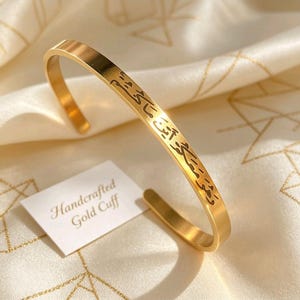 May include: A gold-colored cuff bracelet with engraved text. The bracelet is open and has a smooth, polished finish. A small white card with the text "Handcrafted Gold Cuff" is next to the bracelet.