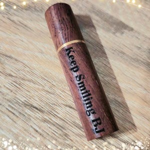 May include: A cylindrical wooden object with the text "Keep Smiling BJ" engraved on its side. The wood has a rich, dark brown color with a visible grain. A thin gold band encircles the object near the top. The object is lying on a wooden surface.