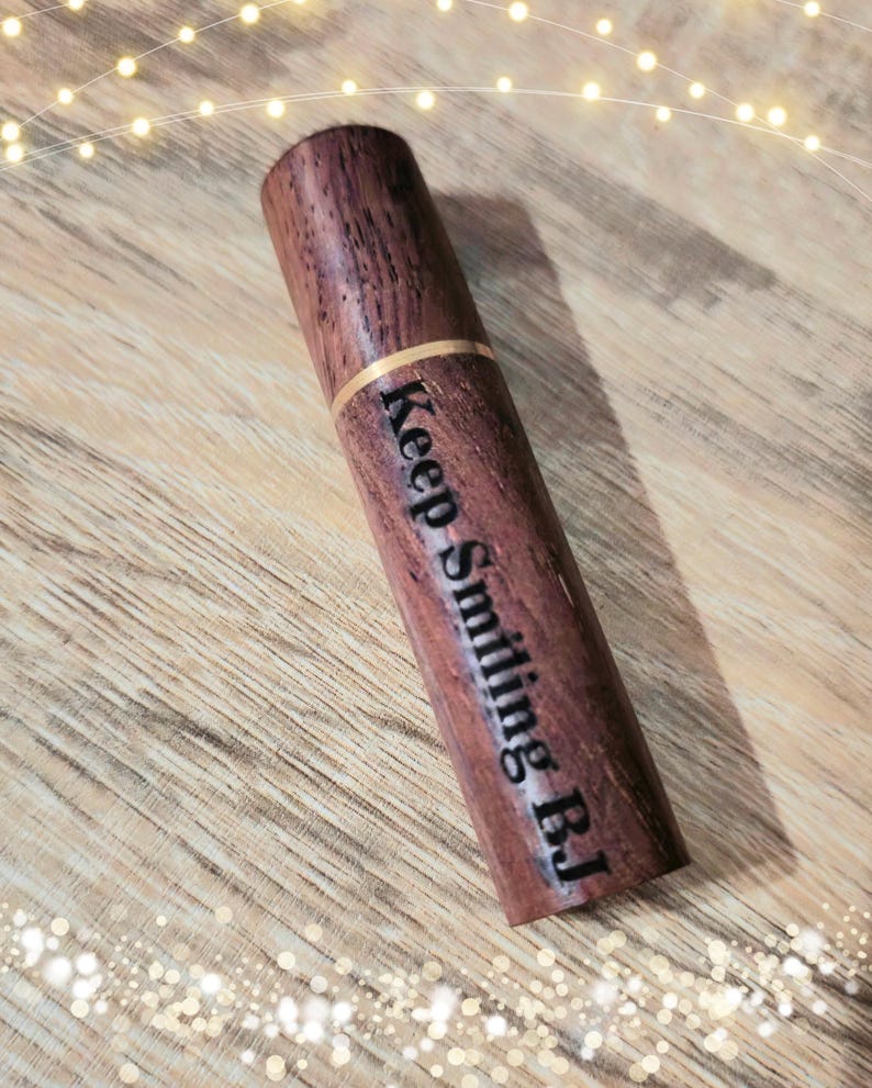 May include: A cylindrical wooden object with the text "Keep Smiling BJ" engraved on its side. The wood has a rich, dark brown color with a visible grain. A thin gold band encircles the object near the top. The object is lying on a wooden surface.
