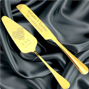 Op de afbeelding: Een goudkleurige taartschep en messenset. De schep heeft een hartvormig ontwerp met Arabisch schrift en de initialen "N & Q" met een datum. Het mes is gegraveerd met de namen "Natalia & Qadar" en een citaat. De set wordt gepresenteerd op een donkere, gedrapeerde stof.