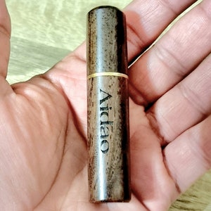 May include: A cylindrical wooden object with the word "Aidao" printed vertically. The object is dark brown with a gold-colored band around the middle. The item is held in an open hand.