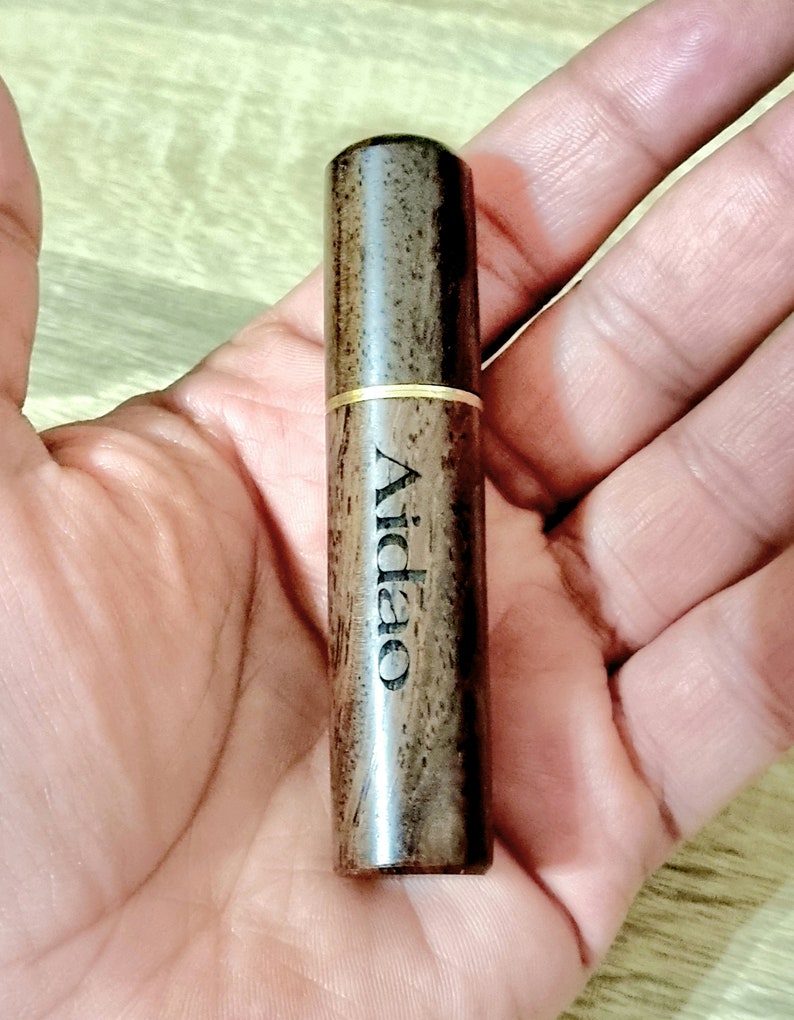 May include: A cylindrical wooden object with the word "Aidao" printed vertically. The object is dark brown with a gold-colored band around the middle. The item is held in an open hand.
