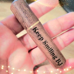 May include: A wooden pen with the words "Keep Smiling BJ" engraved on the side. The pen has a gold band near the top and a dark brown finish. The pen is held in a hand.