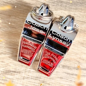 May include: Two silver and red keychains with the text "Coach Jess" and "Coach Lynsay". The keychains are labeled "THE ACME THUNDERER" and "MADE IN ENGLAND". They are on a light brown wooden surface.