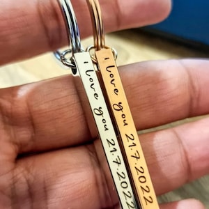 Puede incluir: Dos llaveros, uno plateado y otro dorado, cada uno con un colgante rectangular. Los colgantes están grabados con las palabras "I love you" y la fecha "21.7.2022" en una fuente manuscrita.