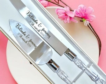 May include: A silver cake knife and server set with clear crystal handles, presented in a white box. The blades are engraved with "Belinda & Jimmy 27th March 1999". Pink flowers and a branch are in the background.