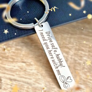 May include: A silver-colored rectangular keychain with the engraved text "Drive safe, hubby! I need you here with me." The keychain also features an infinity symbol and a heart. The keychain is attached to a silver ring.