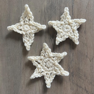 May include: Three handmade crochet stars in off-white with gold metallic thread accents. The stars are arranged on a wooden surface. The stars are a decorative item, possibly for holiday or home decor.