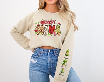 Op de afbeelding: Een beige sweatshirt met een kerstthema-afbeelding waarop "Grinchy Mama" staat met een groen Grinch-personage dat een kerstmanmuts draagt. De mouwen hebben een herhalend patroon van het Grinch-personage.
