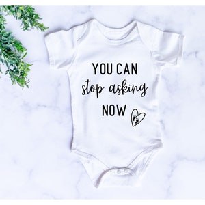 May include: White baby onesie with black text that reads "You can stop asking now" with a small footprint inside a heart.