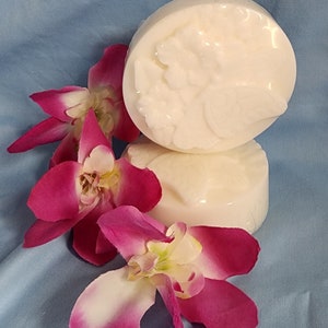 Può includere: Due saponette rotonde bianco sporco con un motivo floreale sono impilate su una superficie azzurra. Due vibranti orchidee rosa e bianche sono disposte vicino ai saponi, aggiungendo un tocco di colore ed eleganza alla composizione.