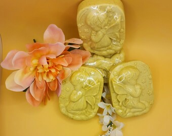May include: Yellow-green soaps with a fairy design are arranged on a yellow surface. The soaps are in a rectangular shape with rounded edges. A large peach-colored flower and small white flowers are placed near the soaps.