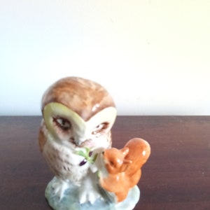 May include: A ceramic figurine of a brown and white owl with a small brown squirrel perched on its back. The owl is holding a green twig in its beak. The figurine is set on a blue and green base.